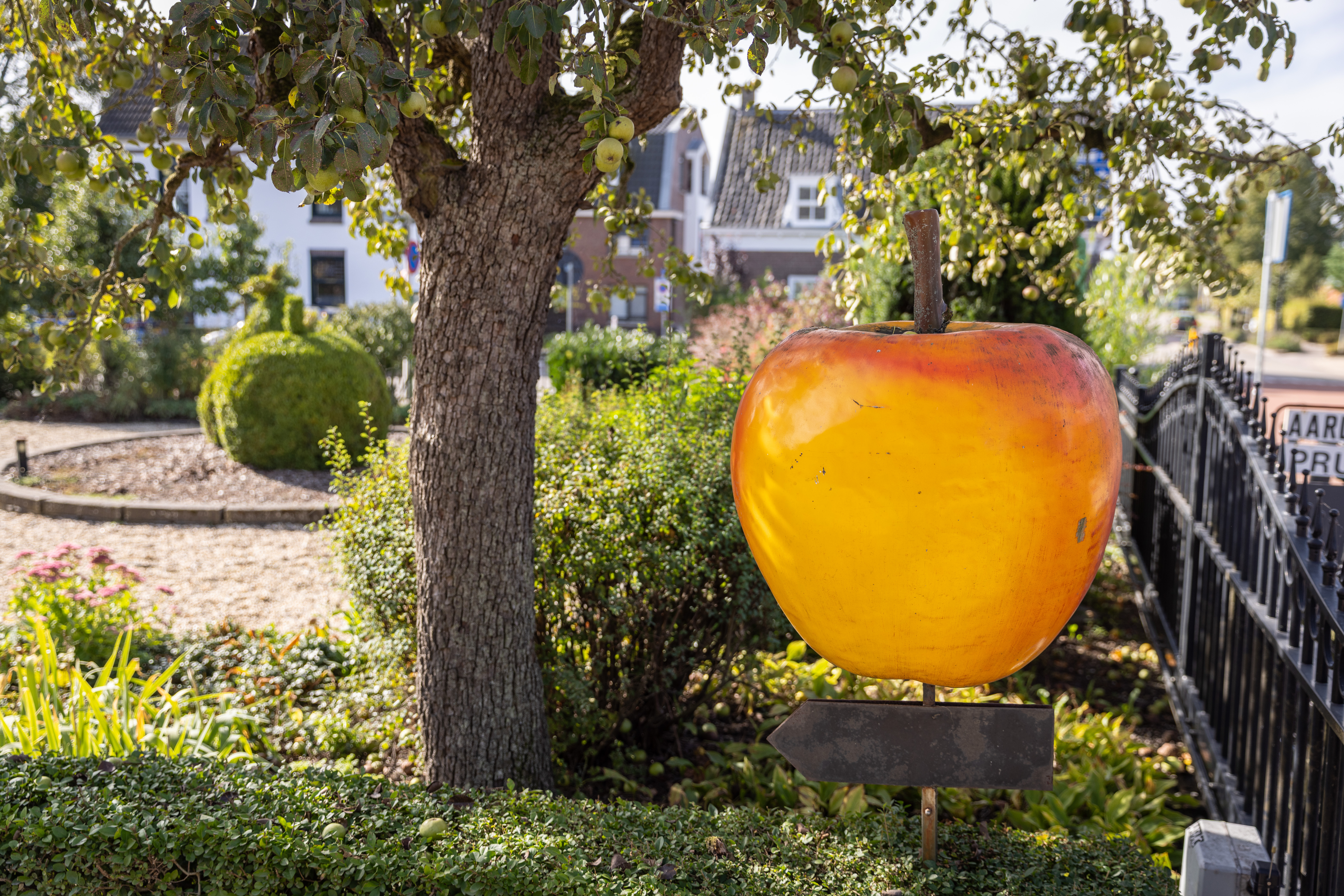 Foto van een tuin met struiken en een boom en een bord van een appel 